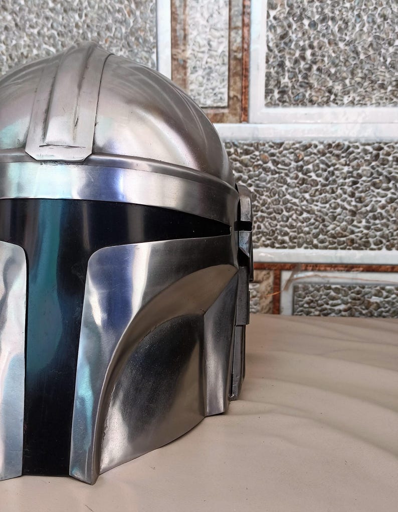May include: A metallic silver and black helmet with a distinctive design. The helmet features a rounded top with vertical ridges and a visor area. The lower section is black, contrasting with the silver upper portion. The helmet is likely a prop or costume piece.