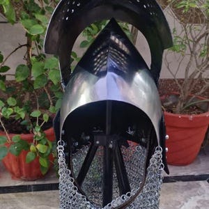 Peut inclure: Casque m&eacute;di&eacute;val en m&eacute;tal poli, avec un sommet conique et une visi&egrave;re. Le casque comprend une protection de cou en cotte de mailles et est expos&eacute; sur un support noir, sur fond de verdure.