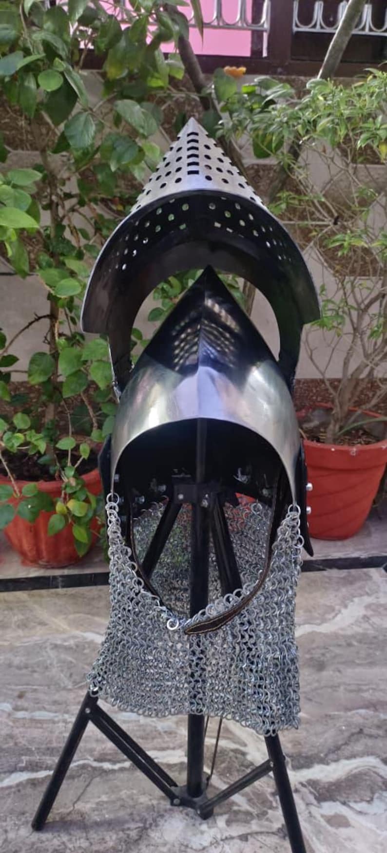 Peut inclure: Casque m&eacute;di&eacute;val en m&eacute;tal poli, avec un sommet conique et une visi&egrave;re. Le casque comprend une protection de cou en cotte de mailles et est expos&eacute; sur un support noir, sur fond de verdure.