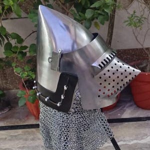 Peut inclure: Casque de style m&eacute;di&eacute;val avec un sommet pointu et une visi&egrave;re &agrave; petits trous. Le casque est en m&eacute;tal brillant et comporte une protection de cou en cotte de mailles. Il poss&egrave;de une section en cuir noir avec des rivets d&eacute;coratifs.