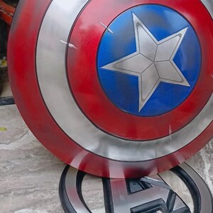 May include: A Captain America shield prop, featuring a red, silver, and blue color scheme. The shield has a circular design with a central blue circle and a white star. The shield is resting on a black and silver Avengers logo.