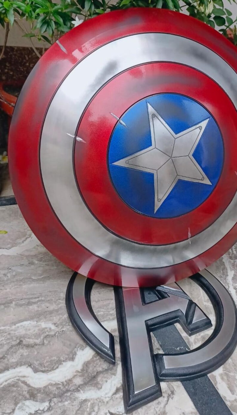 May include: A Captain America shield prop, featuring a red, silver, and blue color scheme. The shield has a circular design with a central blue circle and a white star. The shield is resting on a black and silver Avengers logo.
