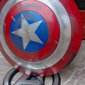 May include: A Captain America shield replica, featuring a red and silver concentric circle design with a blue central disc and a white star. The shield is displayed on a stand with the Avengers logo.