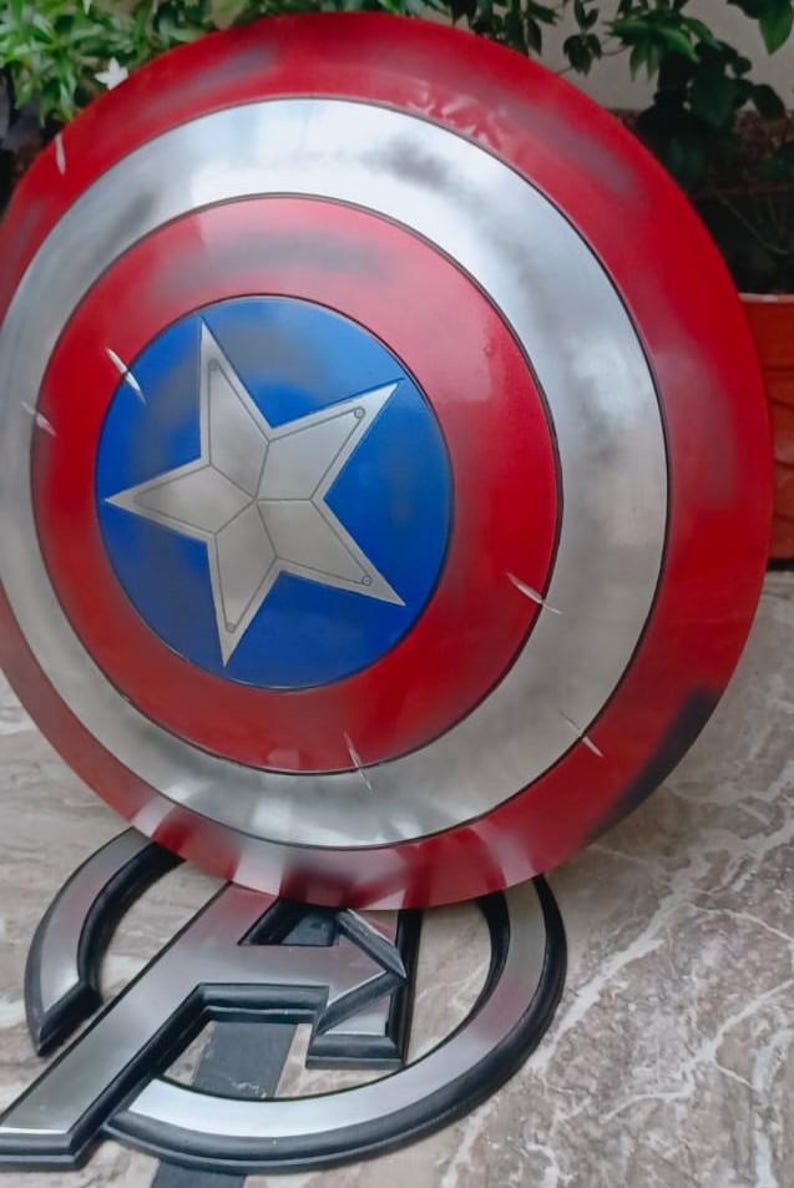 May include: A Captain America shield replica, featuring a red and silver concentric circle design with a blue central disc and a white star. The shield is displayed on a stand with the Avengers logo.