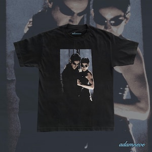 Könnte beinhalten: Ein schwarzes Kurzarm-T-Shirt mit einem rechteckigen Grafikdruck von zwei Figuren, die eine dunkle Sonnenbrille und schwarze Kleidung tragen. Das T-Shirt hat einen Rundhalsausschnitt und den Schriftzug adamneve in Hellblau.