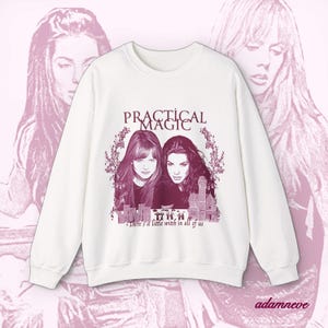 Könnte beinhalten: Ein weißes Sweatshirt mit Rundhalsausschnitt und einem kastanienbraunen Grafikdruck von zwei Frauen, einem Haus sowie dem Text "PRACTICAL MAGIC" und "There's a little witch in all of us". Das Design enthält florale Akzente um das Porträt.