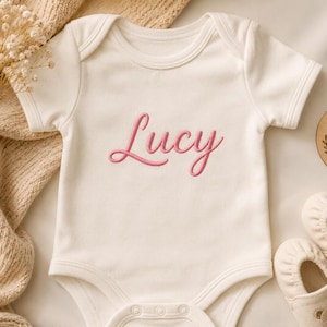 May include: A cream-colored short-sleeve baby bodysuit featuring the name Lucy embroidered in pink script across the chest. The garment includes a round neckline and snap closures at the bottom, accompanied by a pair of small cream-colored baby shoes.