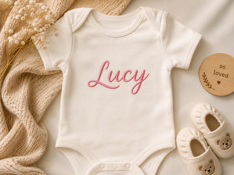 May include: A cream-colored short-sleeve baby bodysuit featuring the name Lucy embroidered in pink script across the chest. The garment includes a round neckline and snap closures at the bottom, accompanied by a pair of small cream-colored baby shoes.