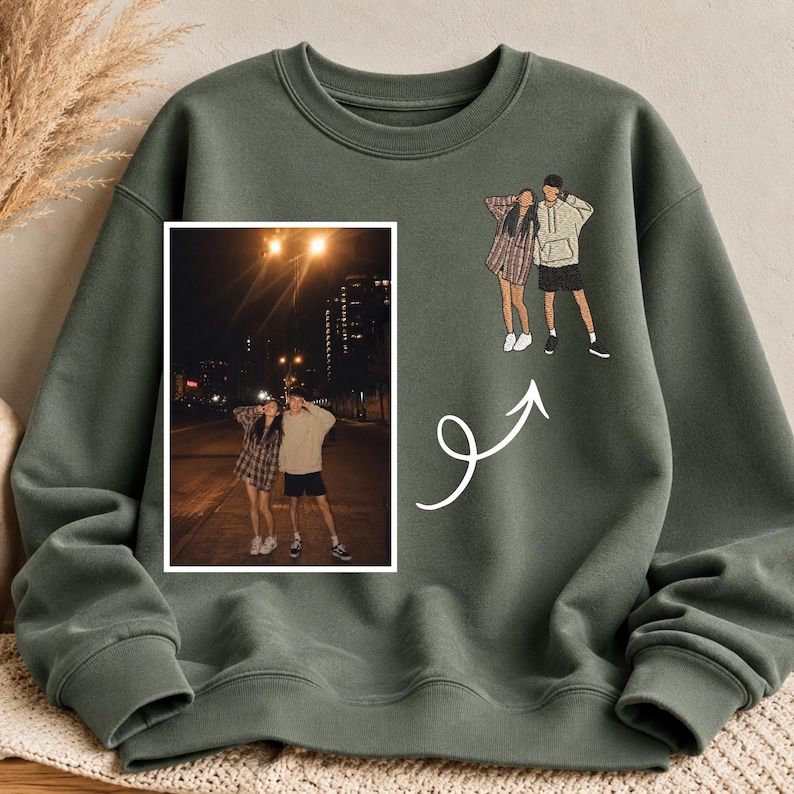May include: An olive green crewneck sweatshirt featuring a rectangular photograph of two people standing on a street at night, alongside a stylized line art illustration of the same two people and a white curved arrow pointing toward the photo.