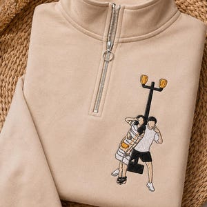 May include: A beige quarter-zip sweatshirt featuring an embroidered graphic of two figures standing by a tall black street lamp. The figures are depicted in casual clothing, with one wearing a striped top and the other a plain white t-shirt and shorts.