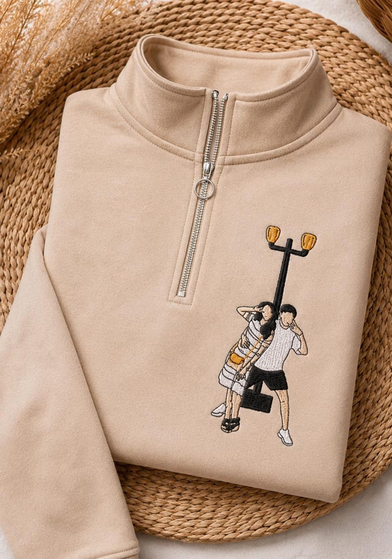 May include: A beige quarter-zip sweatshirt featuring an embroidered graphic of two figures standing by a tall black street lamp. The figures are depicted in casual clothing, with one wearing a striped top and the other a plain white t-shirt and shorts.