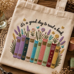 Puede incluir: Un bolso de tela beige con un diseño bordado de libros coloridos alineados, acompañados de flores y pequeñas mariposas que emergen de la parte superior. El texto "it's a good day to read a book" está bordado en hilo negro arriba.