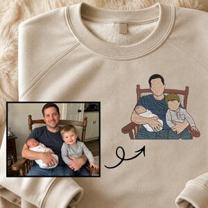 Puede incluir: Sudadera beige con una ilustración bordada de una persona sosteniendo a dos niños. La imagen también incluye una fotografía enmarcada de una persona sosteniendo a dos niños. Una flecha negra apunta de la fotografía a la imagen bordada.