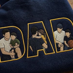 May include: A navy blue sweatshirt featuring the word DAD embroidered in large yellow-outlined letters. Inside each letter, there is a detailed embroidered illustration of a parent with a child, depicting various bonding moments and activities.