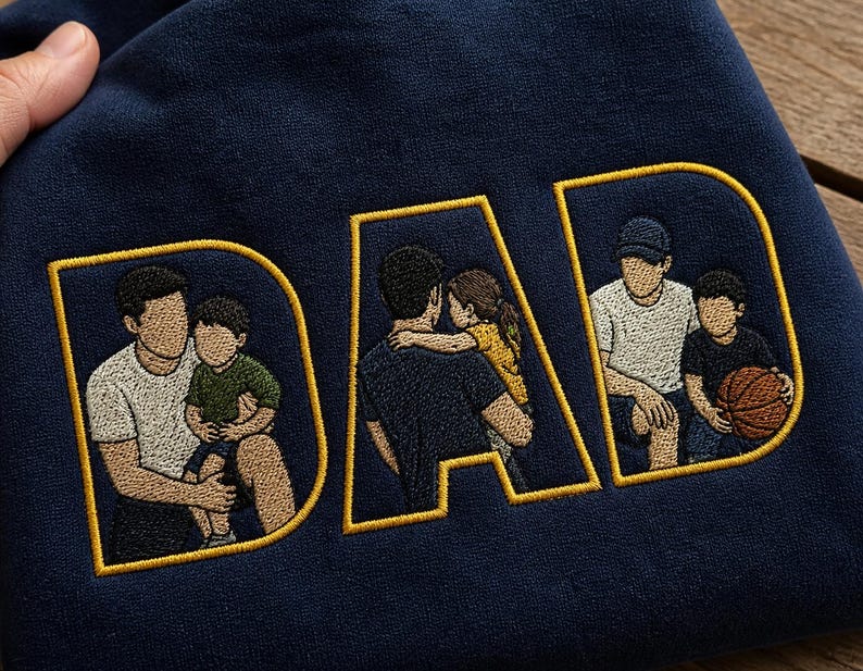 May include: A navy blue sweatshirt featuring the word DAD embroidered in large yellow-outlined letters. Inside each letter, there is a detailed embroidered illustration of a parent with a child, depicting various bonding moments and activities.