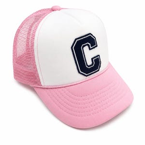 May include: A pink and white trucker hat featuring a large, dark navy blue letter C patch on the front white foam panel. The cap includes a pink mesh back, a pink brim with stitched detailing, and a matching pink button on the top crown.