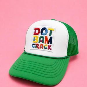 May include: A green and white trucker hat featuring a white front panel with an embroidered patch. The patch displays the text DOT BAM CRACK in multi-colored, block-style letters. The hat has a green brim and a green mesh back panel.