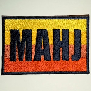 May include: A rectangular embroidered patch featuring the letters MAHJ in bold black thread. The background consists of four horizontal stripes in a gradient of colors, transitioning from bright yellow at the top to orange at the bottom.