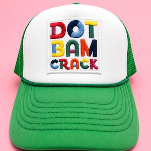 May include: A green and white trucker hat with the embroidered text "DOT BAM CRACK" in colorful letters on a white square patch. The hat has a green brim and mesh back.