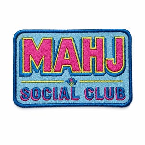 May include: A rectangular embroidered patch with a light blue background and a dark blue border. The text MAHJ is in large pink letters with yellow outlines, and the words SOCIAL CLUB appear below in smaller pink letters with a yellow line separator.