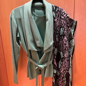 May include: A sage green jacket with a draped collar and a matching tie belt. A patterned scarf with black, teal, and purple snake print is draped over the jacket. The jacket is hanging on a metal hanger.