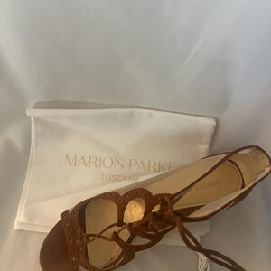 Op de afbeelding: Bruine leren sandalen met een vetersluiting en decoratieve uitsparingen. De sandalen hebben een platte zool en een klein kwastje. Op de achtergrond een witte tas met de tekst "MARION PARKE TOSCANA".