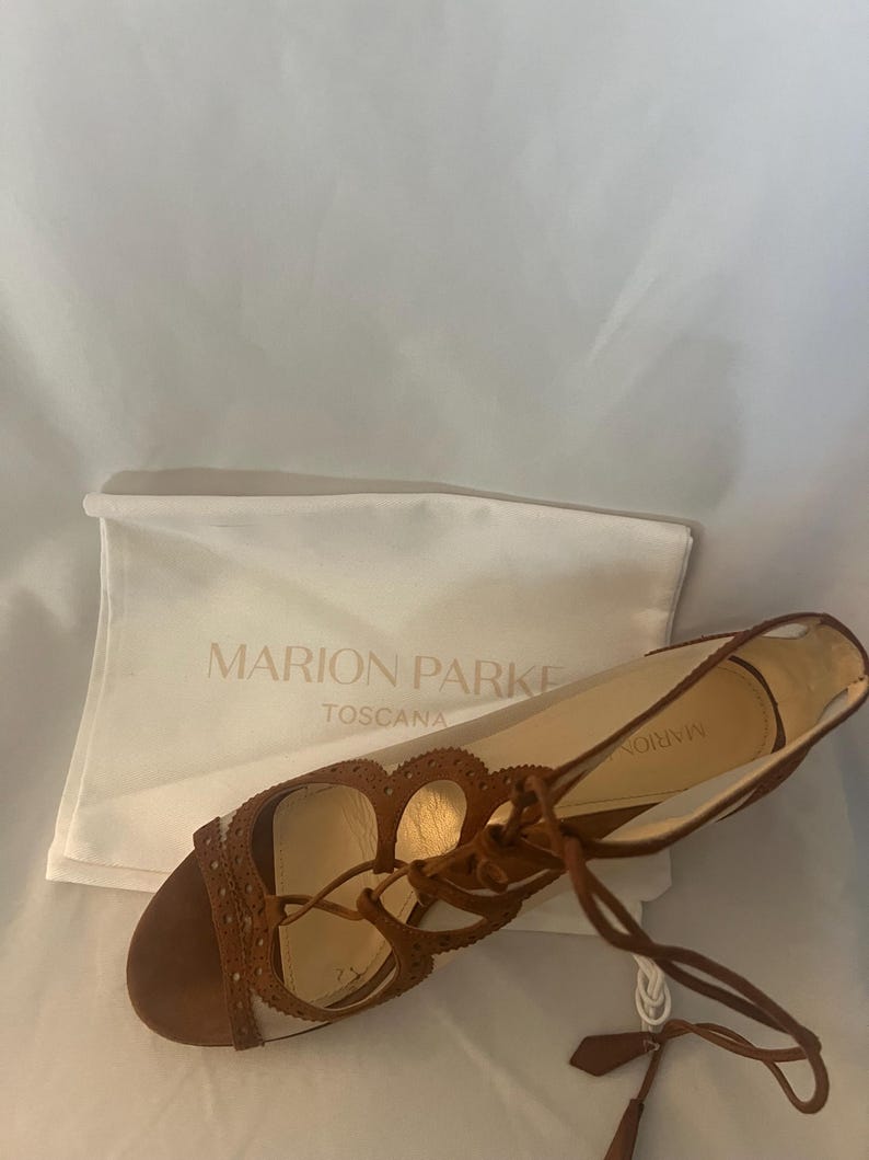 Op de afbeelding: Bruine leren sandalen met een vetersluiting en decoratieve uitsparingen. De sandalen hebben een platte zool en een klein kwastje. Op de achtergrond een witte tas met de tekst "MARION PARKE TOSCANA".