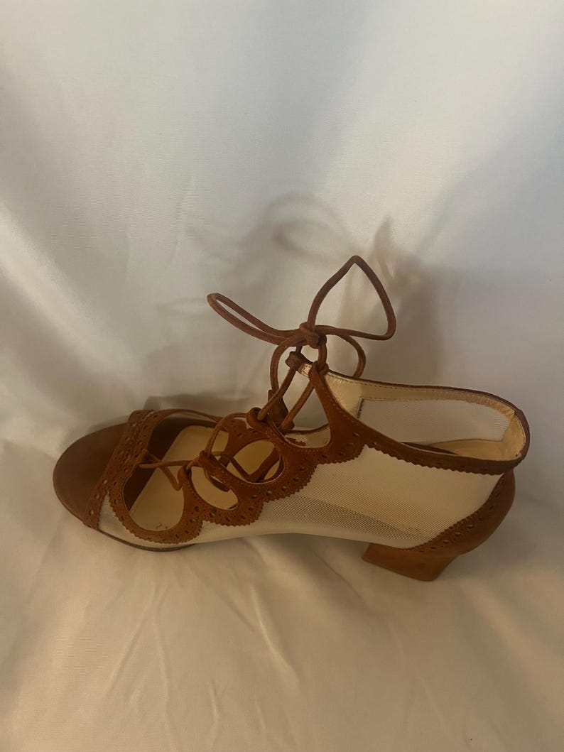 Op de afbeelding: Bruine en cr&egrave;mekleurige vetersandalen. De sandalen hebben een bruine su&egrave;de neus en hiel, met een cr&egrave;mekleurig mesh bovenwerk. Het ontwerp omvat decoratieve uitgesneden details en een kleine hak. De veters zijn bruin.