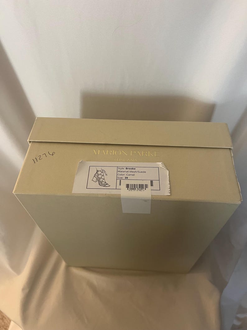 Op de afbeelding: Een beige schoenendoos met de merknaam "MARION PARKE" op de bovenkant. Een sticker aan de zijkant geeft de schoenstijl "Brooke", materiaal "Mesh/Suede", kleur "Camel" en maat "39" weer. De doos staat op een wit oppervlak.