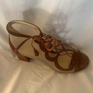 Op de afbeelding: Een bruine su&egrave;de en mesh sandaal met een vetersluiting. De open teen schoen heeft een kleine blokhak en decoratieve uitsparingen langs de randen. De sandaal heeft een neutraal mesh bovenwerk en bruine su&egrave;de accenten.