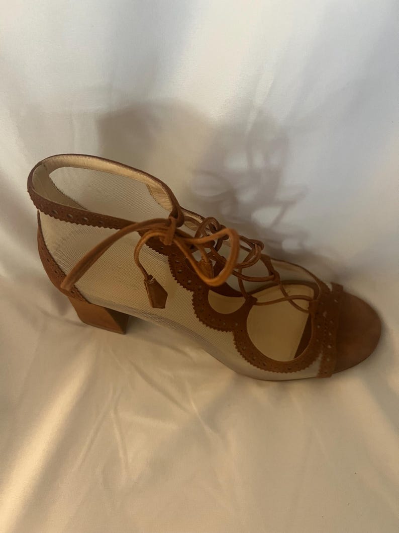 Op de afbeelding: Een bruine su&egrave;de en mesh sandaal met een vetersluiting. De open teen schoen heeft een kleine blokhak en decoratieve uitsparingen langs de randen. De sandaal heeft een neutraal mesh bovenwerk en bruine su&egrave;de accenten.