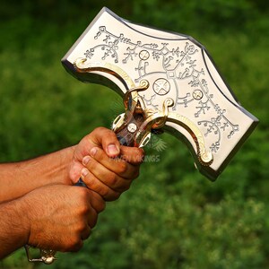 May include: A silver and gold-colored hammer with intricate engraved designs. The hammer's head features detailed patterns and symbols, with gold accents on the edges and handle. The handle is dark brown with a blue grip.