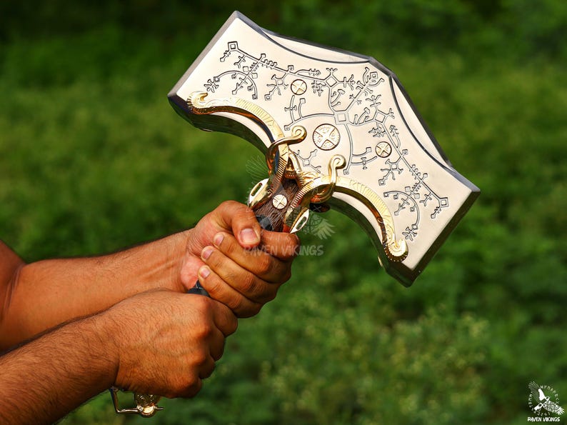 May include: A silver and gold-colored hammer with intricate engraved designs. The hammer's head features detailed patterns and symbols, with gold accents on the edges and handle. The handle is dark brown with a blue grip.