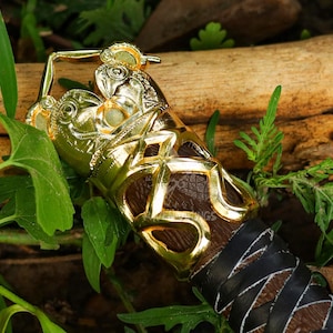 May include: Close-up of a gold-colored decorative object with intricate detailing, possibly a handle or decorative element. The object has a brown leather-like section and is surrounded by green foliage and a wooden log.