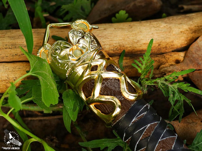 May include: Close-up of a gold-colored decorative object with intricate detailing, possibly a handle or decorative element. The object has a brown leather-like section and is surrounded by green foliage and a wooden log.