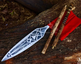 May include: A silver spearhead with black geometric designs, accompanied by two wooden shafts and a red cloth. The spearhead is approximately 20 inches long. The shafts have a wood grain pattern and brass accents. The items are displayed on a wooden surface.