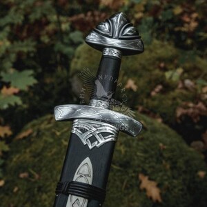 May include: A close-up of a Viking sword with a silver-colored hilt and a black handle. The sword has intricate Celtic knot designs on the hilt and a triangular pommel. The background is blurred, with green and brown foliage.