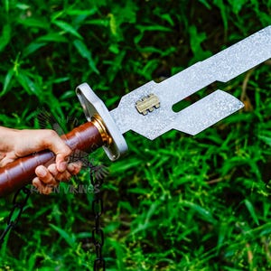 May include: A silver-colored sword with a brown leather handle and gold accents. The blade has a unique cutout design and a small gold emblem. The sword is held against a backdrop of green foliage, suggesting an outdoor setting.