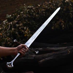 May include: A silver-colored sword with a long, pointed blade. The hilt is silver with a curved design and a black grip. The sword is held in a hand against a backdrop of dark wood and foliage. The text "Raven Vikings" is visible on the hilt.