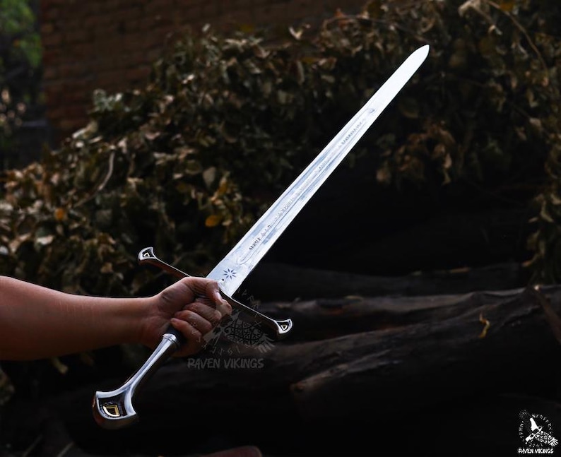 May include: A silver-colored sword with a long, pointed blade. The hilt is silver with a curved design and a black grip. The sword is held in a hand against a backdrop of dark wood and foliage. The text "Raven Vikings" is visible on the hilt.