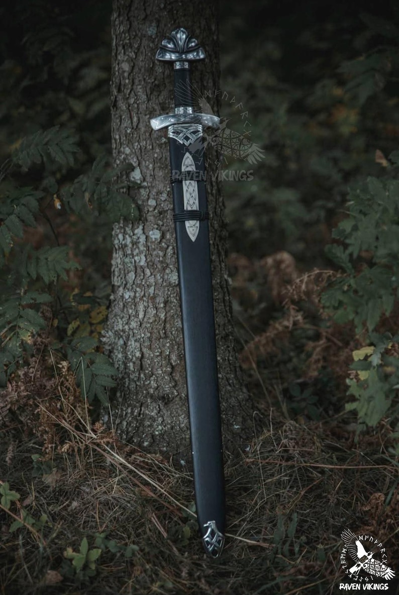 May include: A black sword with a silver handle and accents, resting against a tree trunk. The sword is in a black sheath with silver detailing. The image is set in a forest environment.