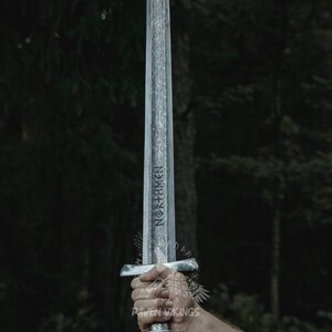 May include: A silver-colored sword with intricate engravings and the word "NORTHMEN" etched on the blade. The sword has a detailed hilt and is held against a blurred forest background.