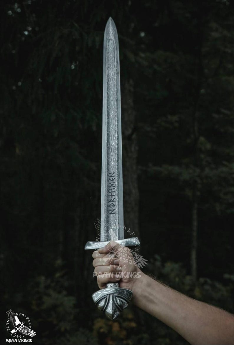 May include: A silver-colored sword with intricate engravings and the word "NORTHMEN" etched on the blade. The sword has a detailed hilt and is held against a blurred forest background.