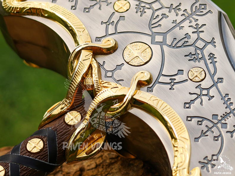 May include: Close-up of a decorative axe head with intricate gold and silver detailing. The axe features a wooden handle wrapped in leather and adorned with gold accents. The blade has etched patterns and gold circular embellishments.