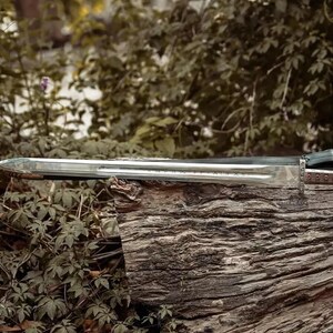 May include: A long, silver sword with a detailed handle and a black sheath rests on a weathered log. The blade is sharp and reflective, with a decorative handle. The sword is a historical or fantasy item.