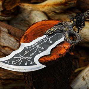 May include: A detailed, decorative knife with a skull-shaped handle and a blade featuring intricate patterns and runes. The handle is gold-colored, and the blade is silver and black. The knife rests on a wooden surface, showcasing its craftsmanship.