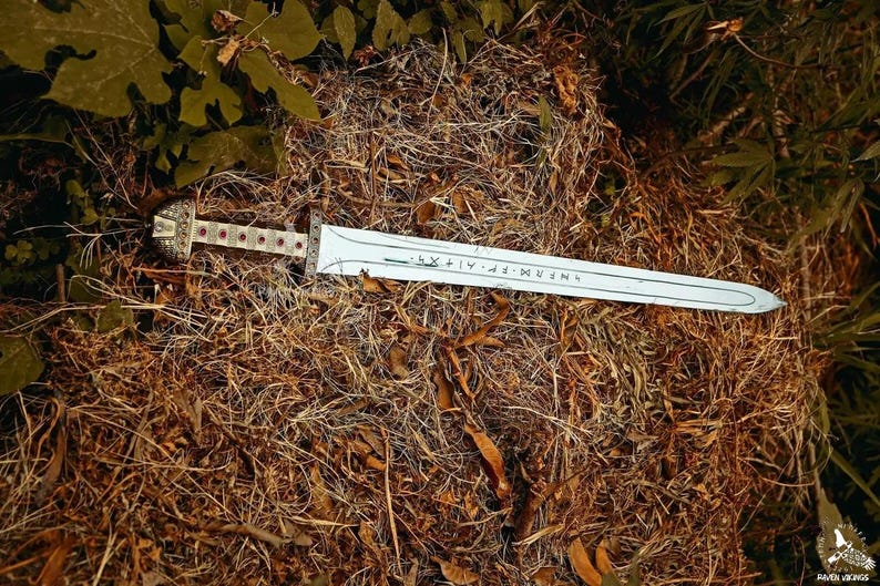 May include: A decorative sword with a silver blade and ornate handle, resting on a bed of dry grass and leaves. The handle features red accents. The blade has engraved text.