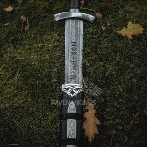 May include: A detailed image of a Viking sword with a silver hilt and a dark wooden handle. The blade is etched with the word "NORTHMEN". The sword is sheathed in a black scabbard with silver accents. The background is a bed of green moss.