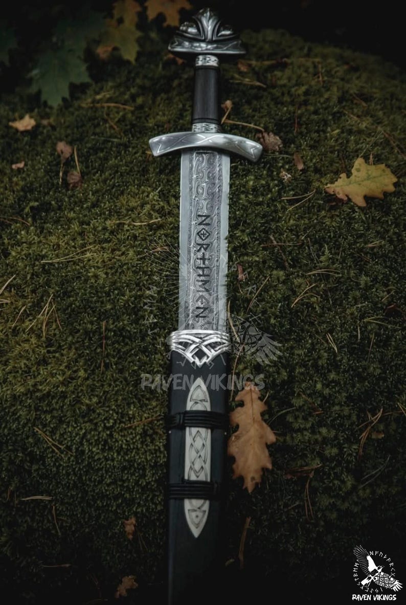May include: A detailed image of a Viking sword with a silver hilt and a dark wooden handle. The blade is etched with the word "NORTHMEN". The sword is sheathed in a black scabbard with silver accents. The background is a bed of green moss.