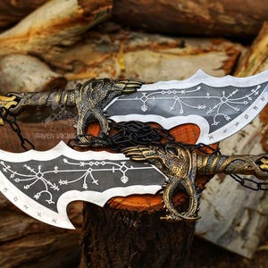 May include: Two silver and black blades with ornate gold handles and chains. The blades have a curved, serrated edge and are decorated with white symbols. The handles feature dragon-like figures. The blades are displayed on a wooden surface.